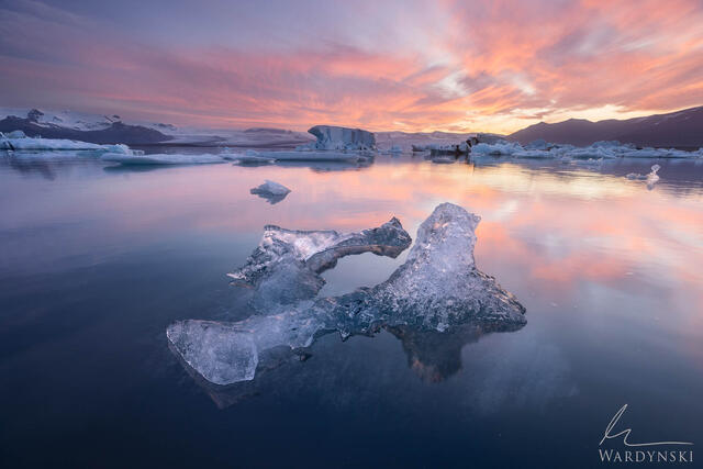 This is contemporary landscape photograph of a large fragment of glacier ice floats in the Jökulsárlón Glacier Lagoon at sunset.