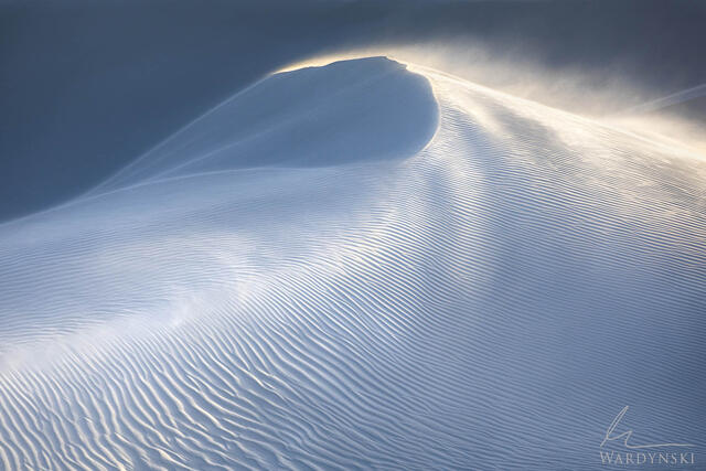 Backlit sand blowing off the top of the dunes in White Sands National Park.
